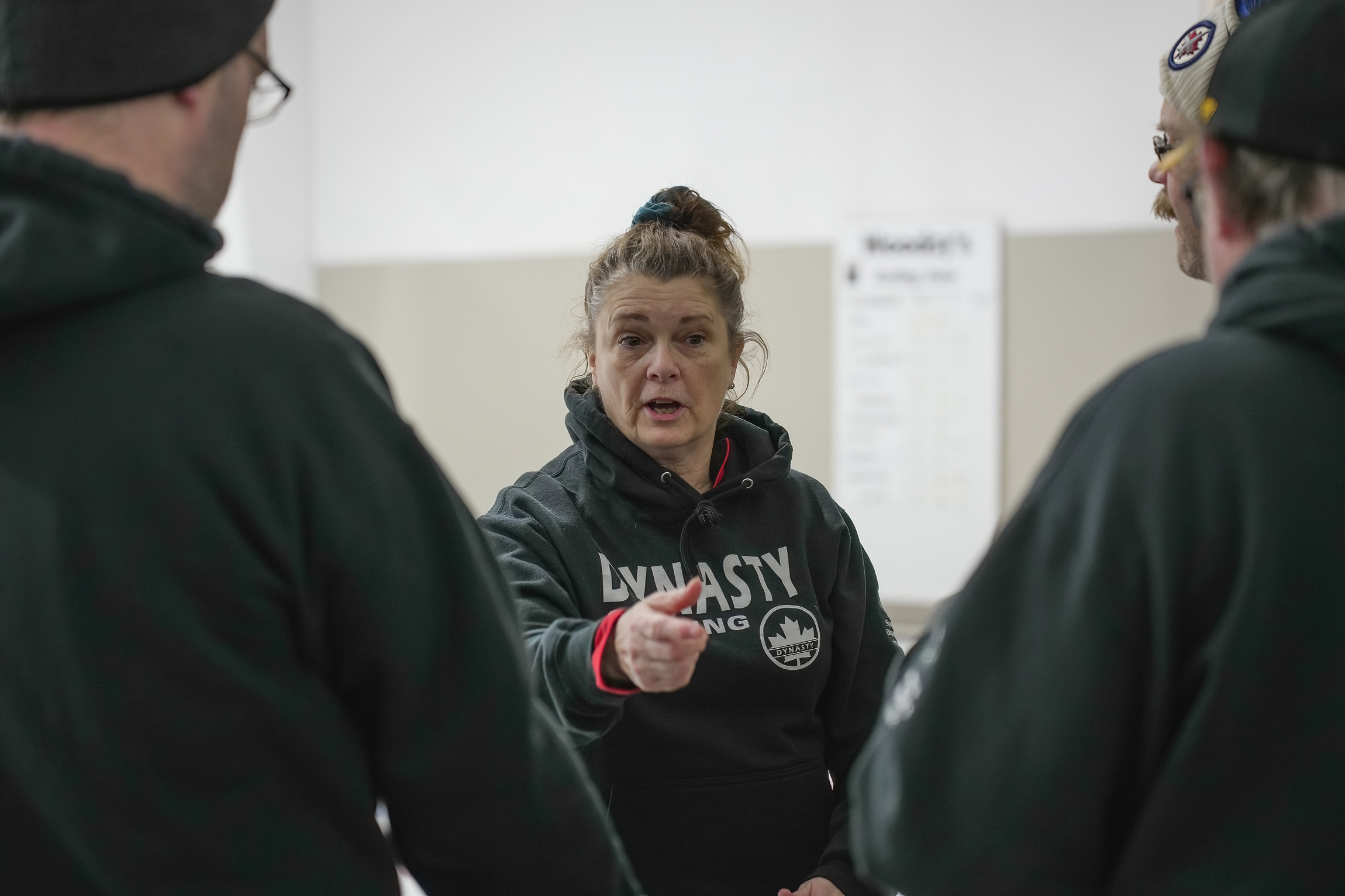 A coach, coaching her team. She is in the middle of speaking and pointing at an athlete. Two athletes can be seen in front of her, but they have their backs to the camera and are out of focus. A coach, coaching her team. She is in the middle of speaking and pointing at an athlete. Two athletes can be seen in front of her, but they have their backs to the camera and are out of focus.