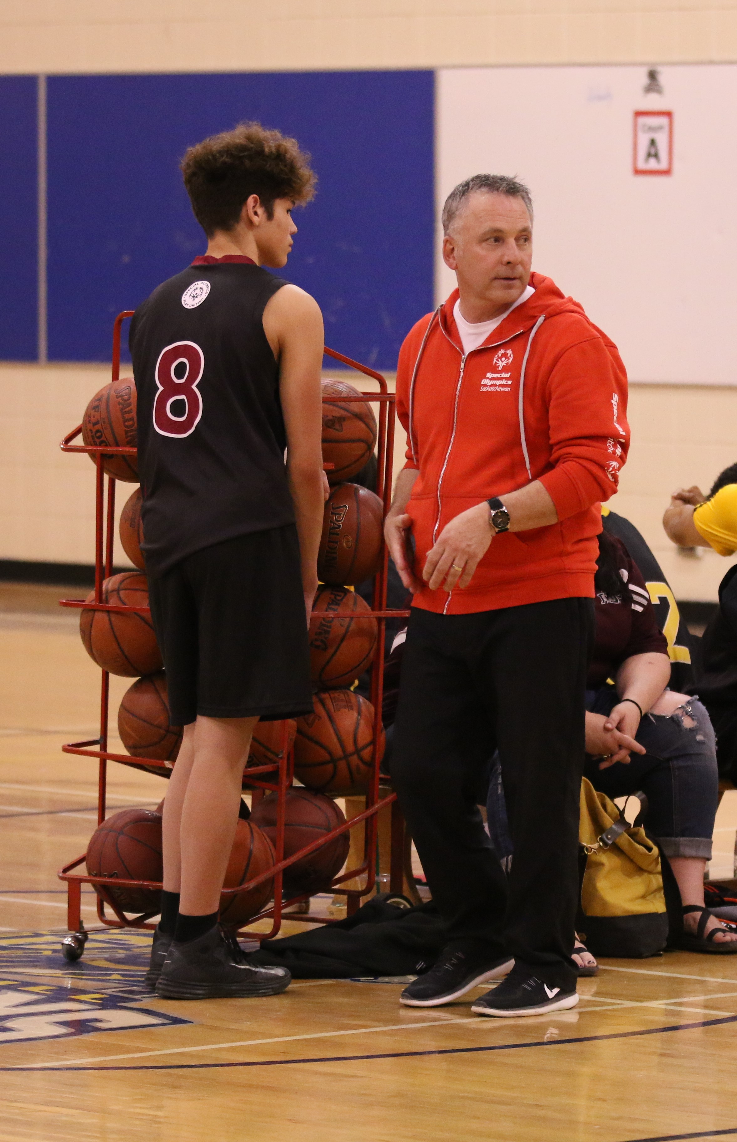 A basketball coach talking to a basketball athlete. They are standing to the side of the court next to a rack filled with basketballs. A basketball coach talking to a basketball athlete. They are standing to the side of the court next to a rack filled with basketballs.