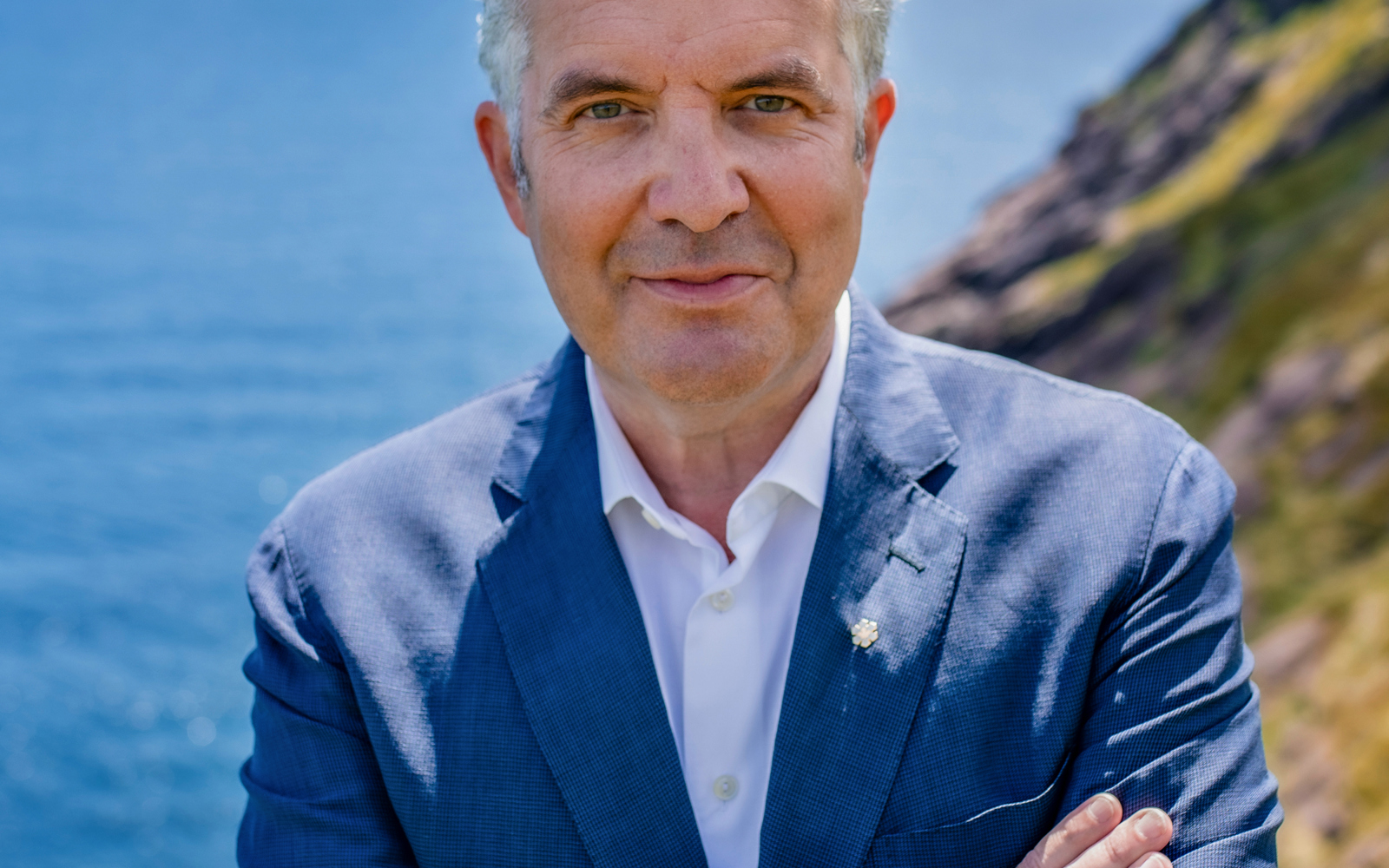 Man in a blue blazer with his arms crossed. Behind him is the ocean.