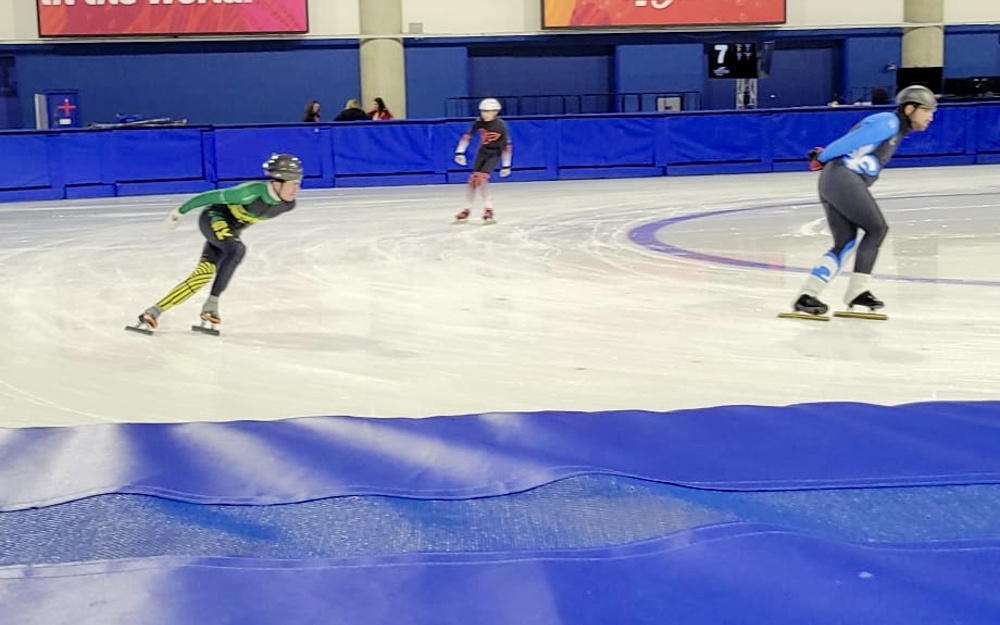 An action shot photo of speed skaters on the ice. Three speed skaters can be seen going around the edge of the rink. They are slightly out of focus due to the speed they are skating.