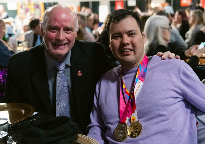 Two individuals are smiling for the camera, sitting at a fancy table. The man on the left is wearing a fancy suit while the athlete on the right is wearing colourful medals around his neck.