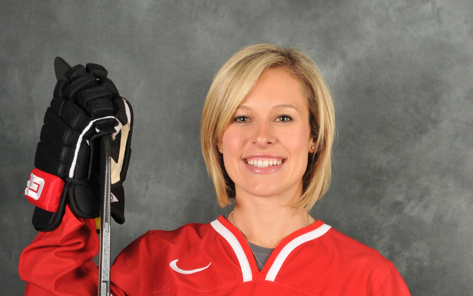 Headshot of a woman short blond hair wearing a Team Canada hockey jersey. She is wearing hockey gloves and holding a hockey stick.