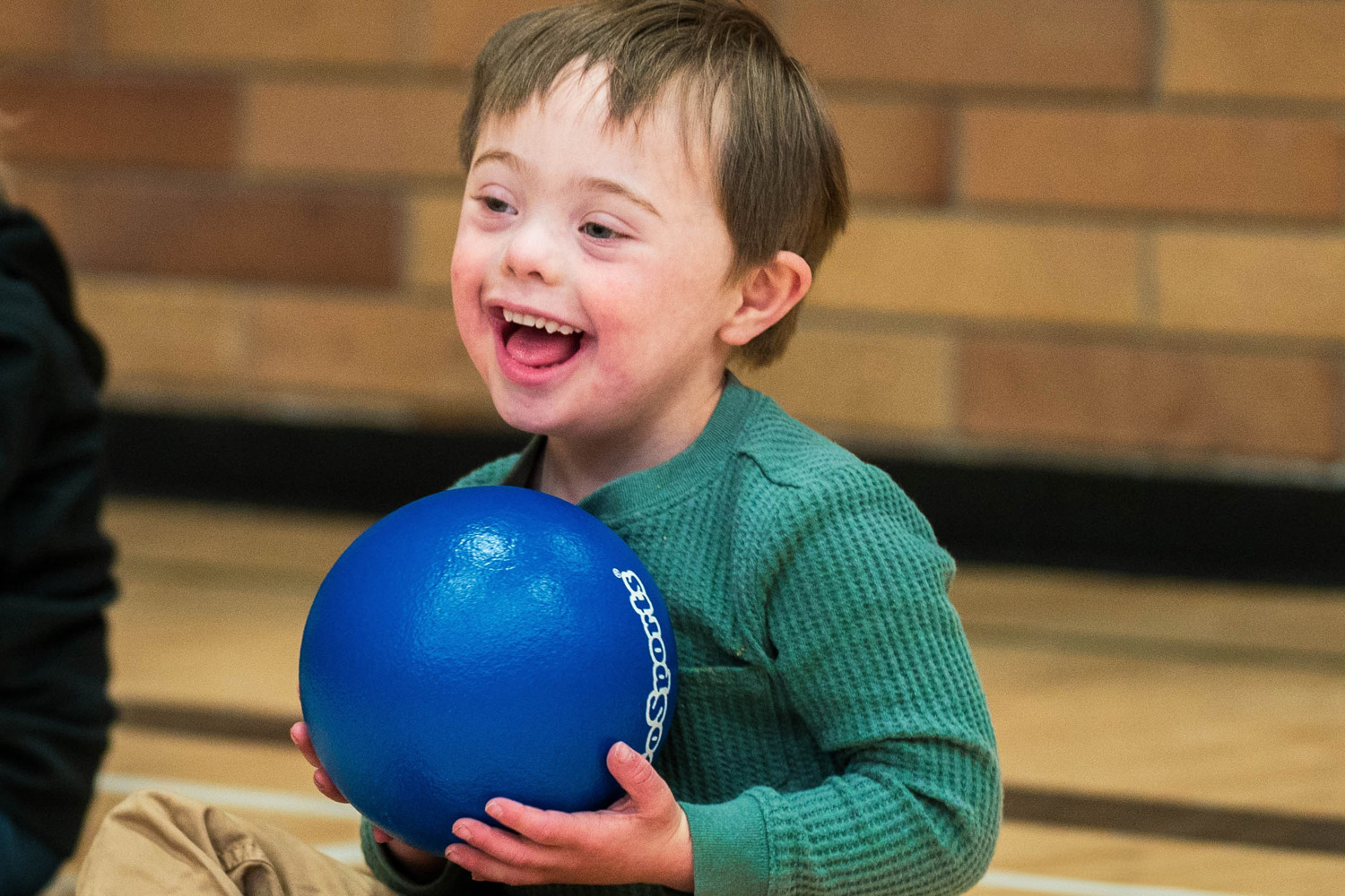 A young boy with intellectual disabilities sits on the gym floor next to another child, smiling while holding a blue rubber ball.