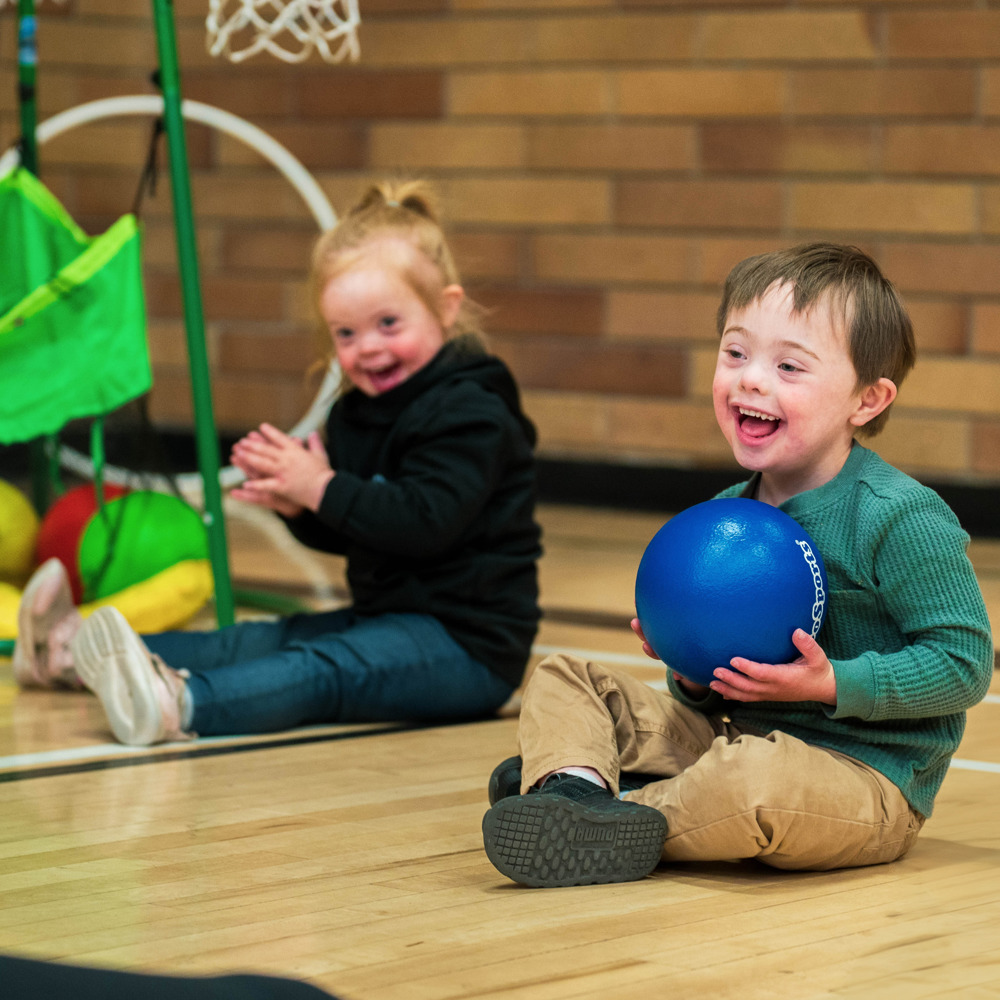 A young boy with intellectual disabilities sits on the gym floor next to another child, smiling while holding a blue rubber ball.