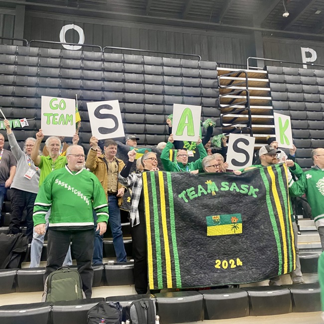 A crowd of people in green, are sitting on the bleachers cheering for Team Saskatchewan. They are holding up flags, banners, and poster boards showing support for the team. Everyone looks happy and are cheering the athletes on.