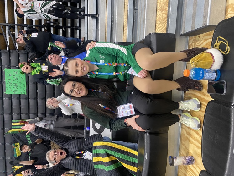 An athlete is sitting with her coach. Their arms are wrapped around behind each other. They are sitting in the bleachers of a sport event. The people behind them are all cheering.