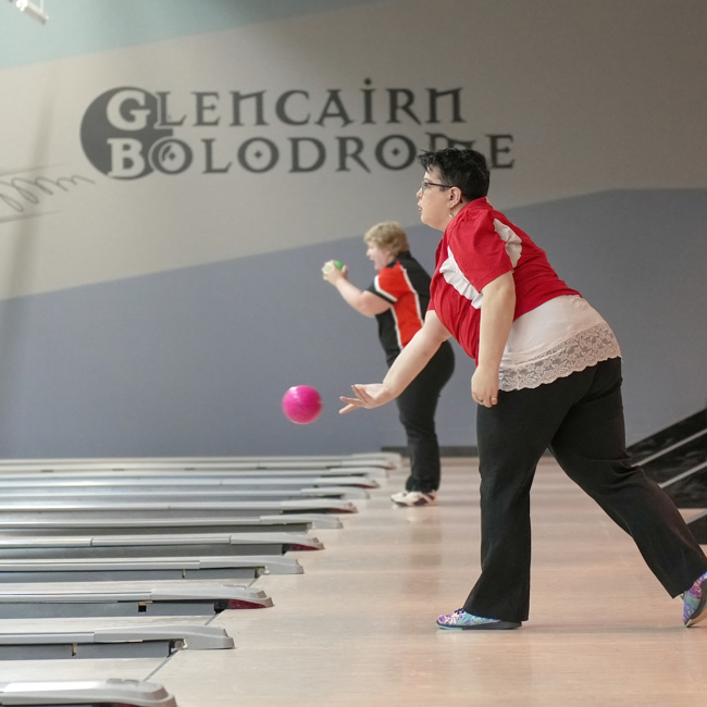 An action shot photo of a bowling athlete. The photo is taken from the side and you can see the start of many different bowling lanes. In the photo, two bowlers can be seen playing the game. The bowler closest to the camera is throwing a pink bowling ball which can be seen midair.