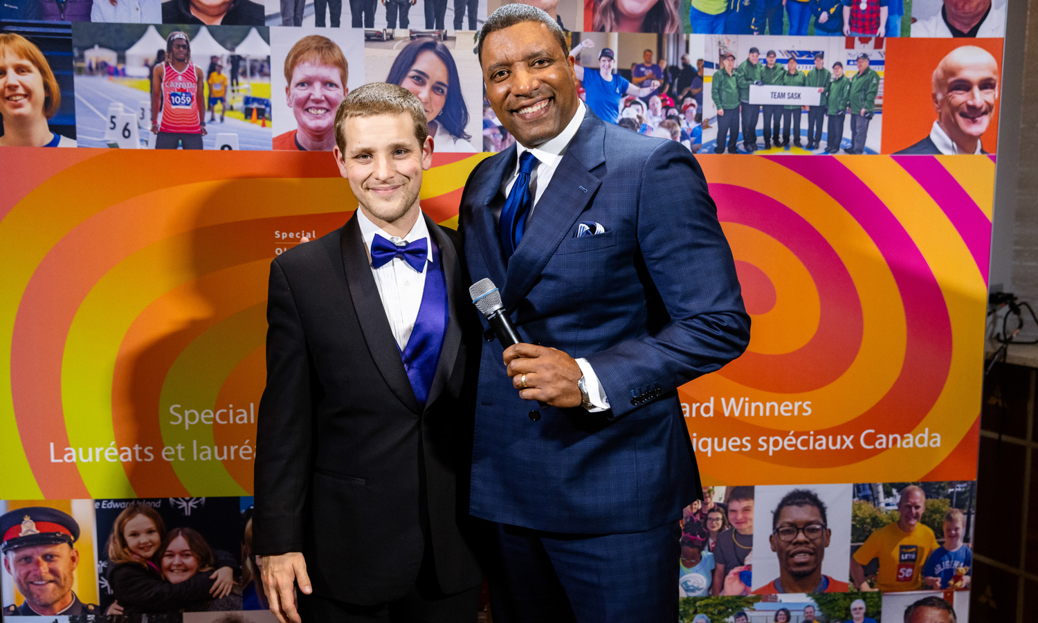 Two men in suits smiling at the camera. One has a microphone in his hand