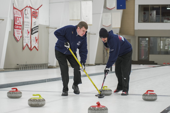 Two athletes participating in a curling match. There are three red and two yellow curling stones dispersed across the ice. The two athletes are using their curling sticks to allow one of the yellow curling stones to move easier.