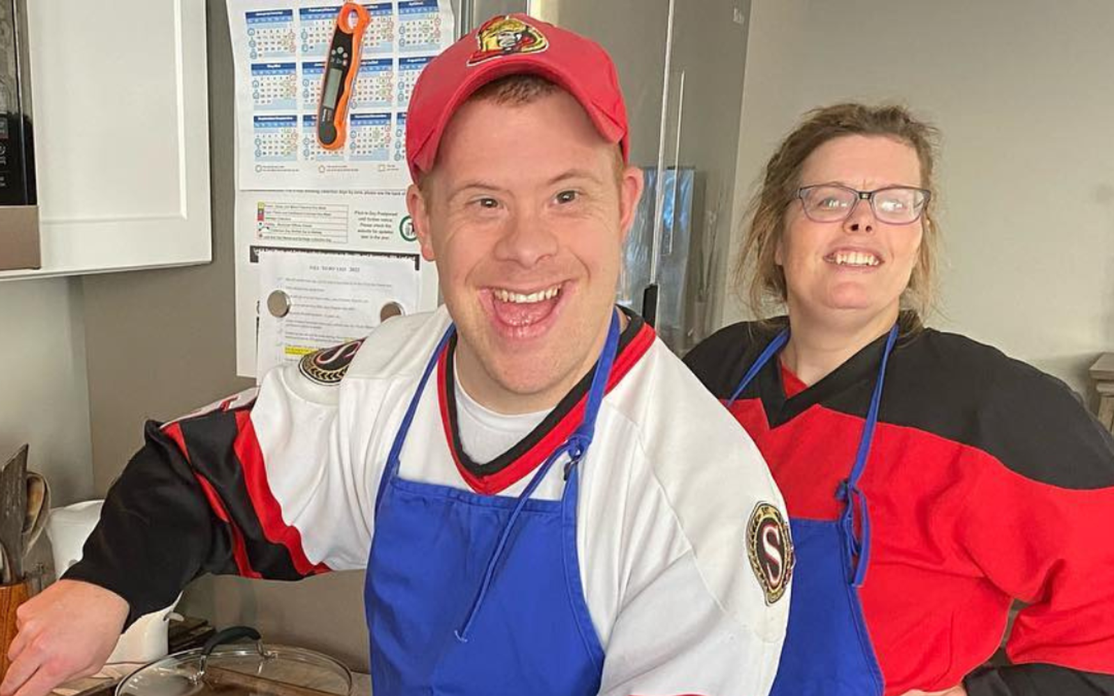 Person wearing a red hat and a blue apron stirring a pot of tomato sauce while another person in a red shirt and blue apron looks on.
