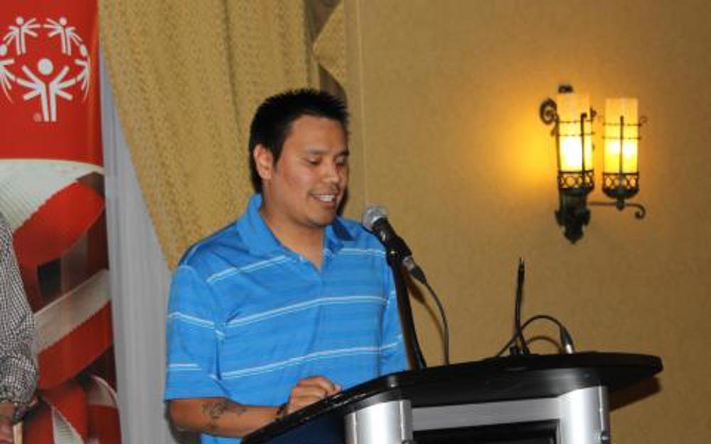 An athlete in a blue striped t-shit is standing at a podium giving a speech.