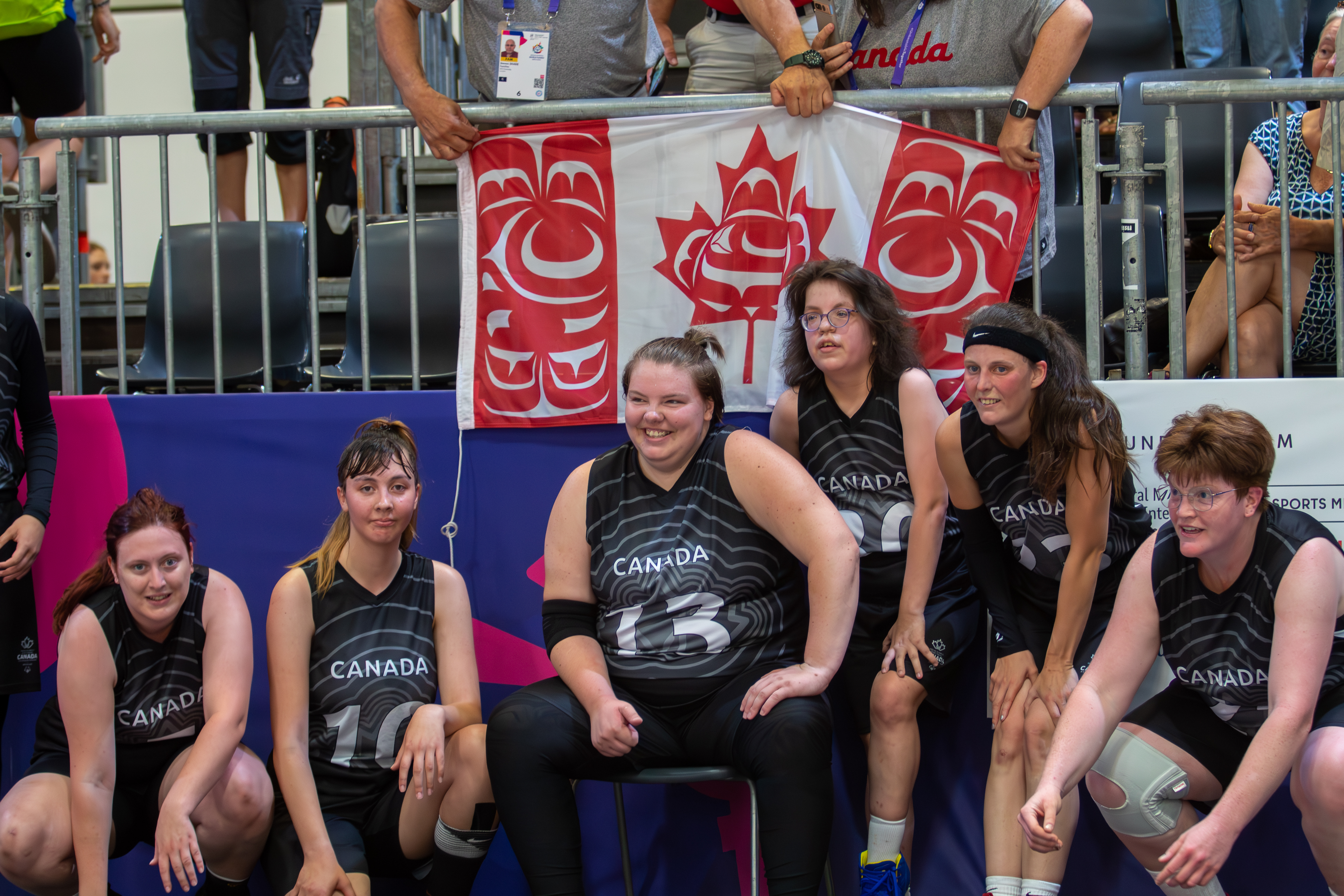 Six Special Olympics Saskatchewan athletes are sitting on the sidelines waiting for their turn to play. They are wearing black Canada jerseys with a different number on each of them. Supporters in the stands behind them are holding a Canadian flag. Six Special Olympics Saskatchewan athletes are sitting on the sidelines waiting for their turn to play. They are wearing black Canada jerseys with a different number on each of them. Supporters in the stands behind them are holding a Canadian flag.