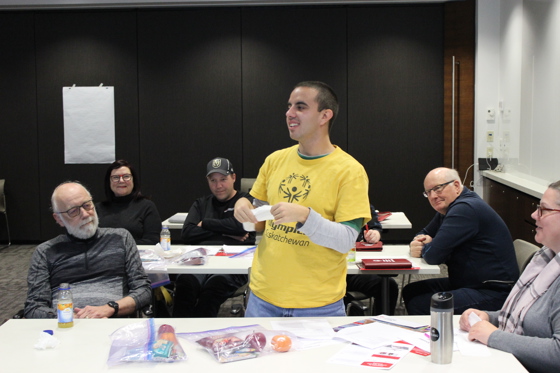 A conference room with three rectangular tables. People can be seen sitting at these three tables. One individual, in the middle of the image, in a yellow shirt, is standing up and presenting something to his peers. Everyone around him is listening intently.