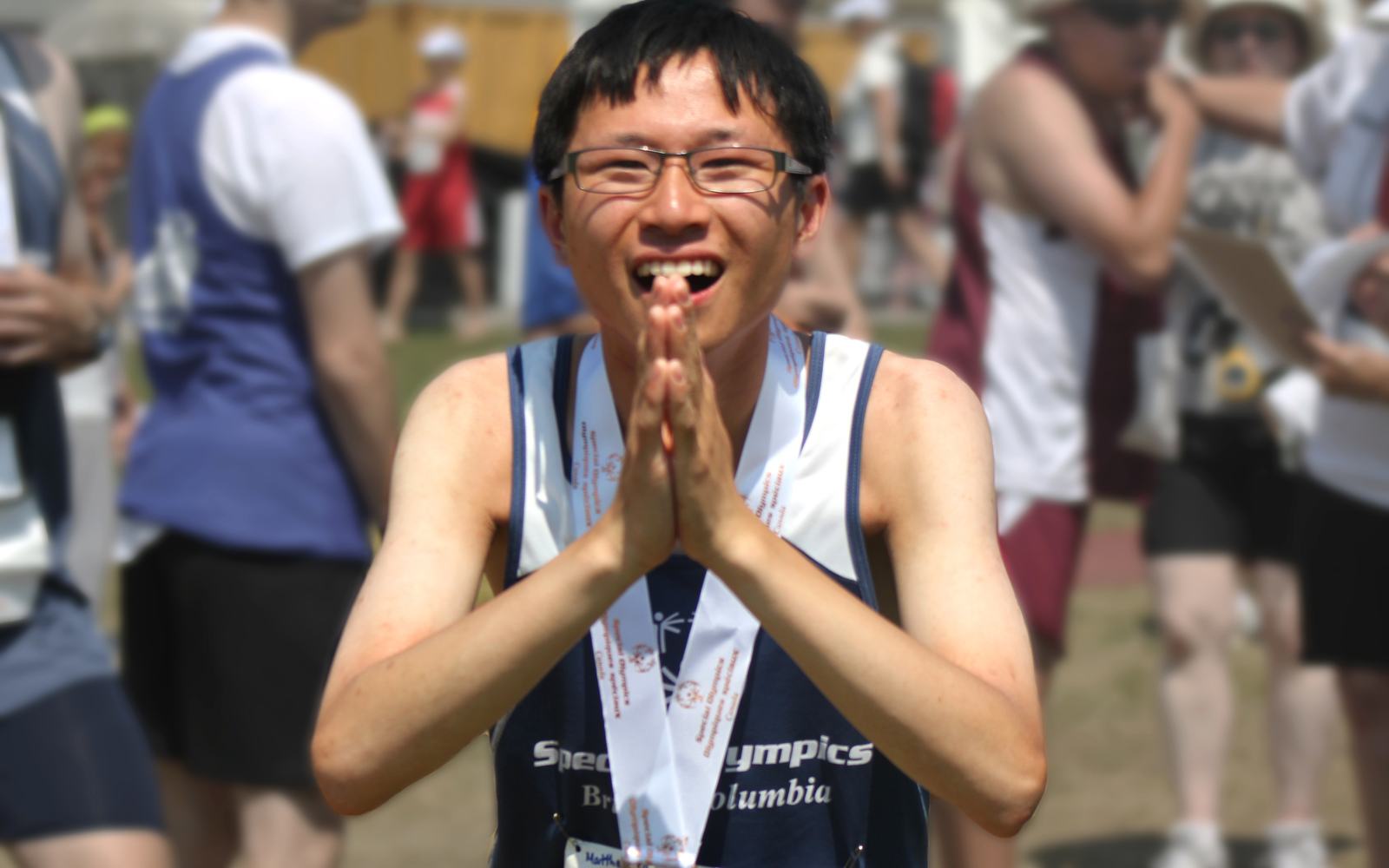 Special Olympics BC athlete with a medal around his neck, looks directly to the camera with hands clasped in front to say thank you.