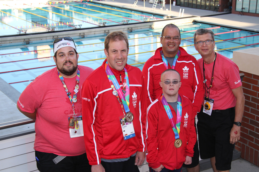 Bill Smith and teammates standing in-front of a swimming pool, showing off their medals. 