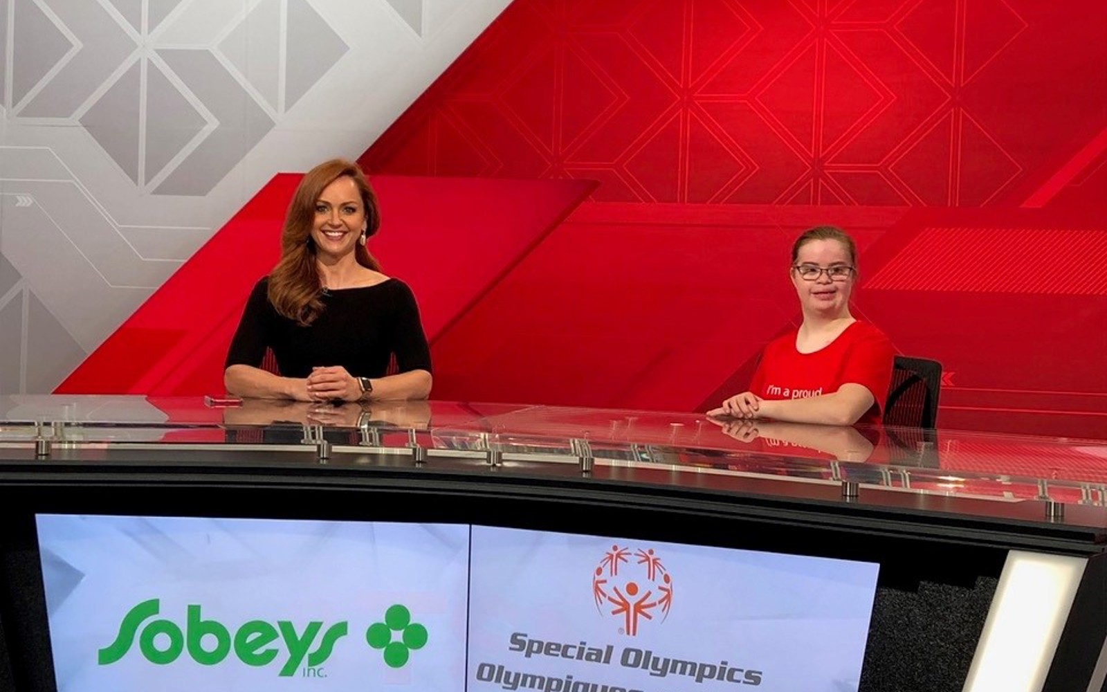 Two women sit behind a desk with the Sobeys logo and the Special Olympics Canada logo on a screen in front of them.