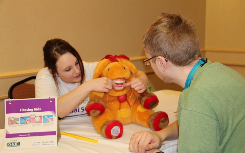 A Healthy Athletes practitioner is demonstrating how to floss your teeth on a stuffed animal with teeth. A athlete in a green t-shirt can be seen from behind watching the demonstration.