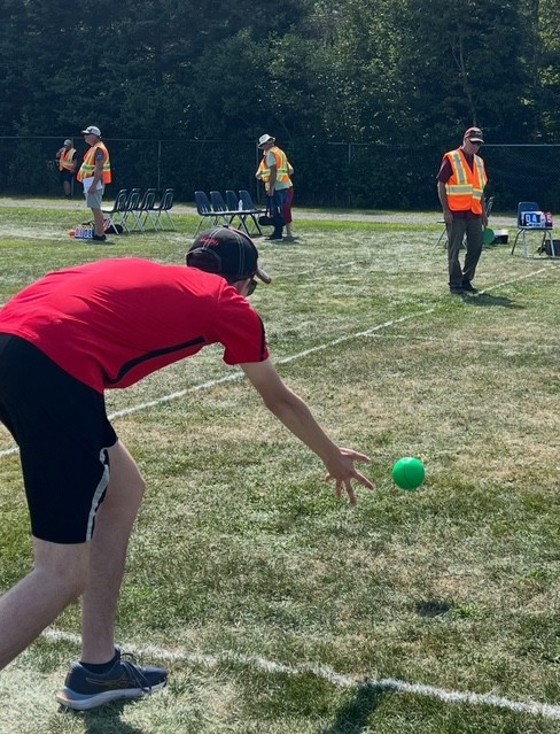 Man in black shorts and red shirt throwing a bocce ball on green grass 