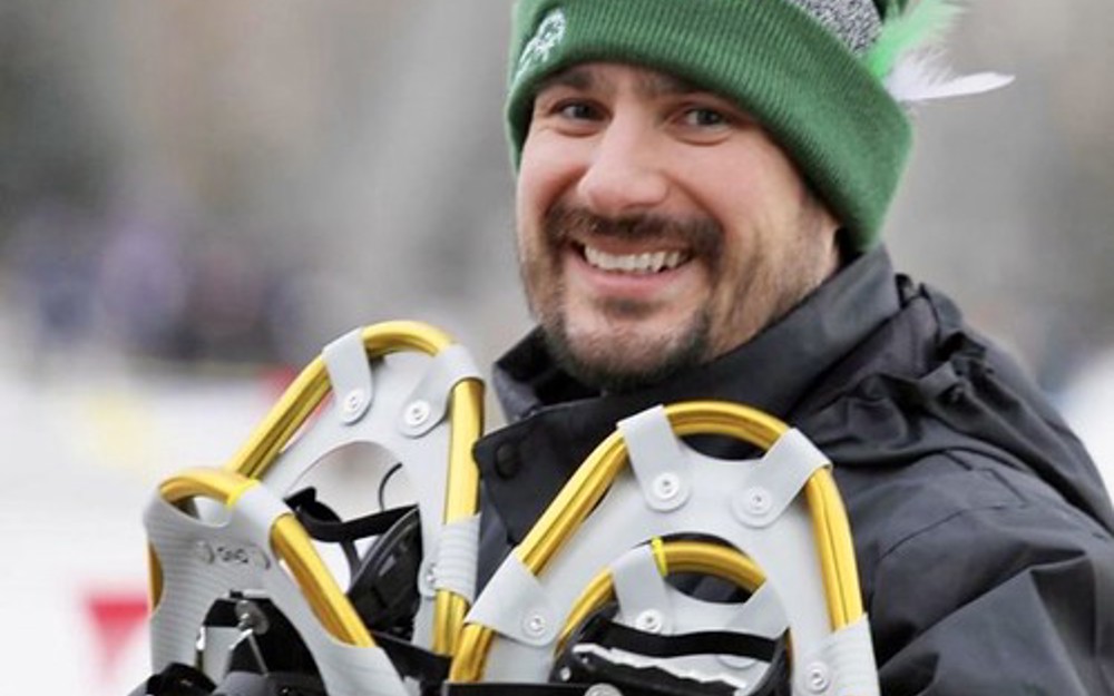 A close up image of Eric, a Special Olympics Saskatchewan coach. He is smiling for the camera and holding snowshoes in his hands. The background behind him is blurred.