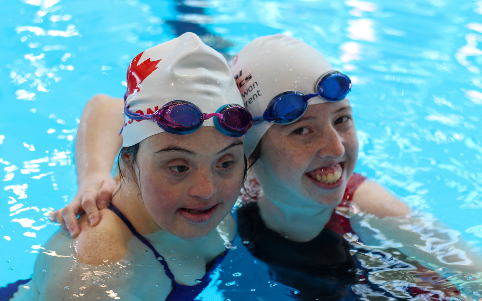 Two girls in a swimming pool. Both are wearing swim caps and have googles on their heads. One is smiling at the camera and the other is looking down at the pool.