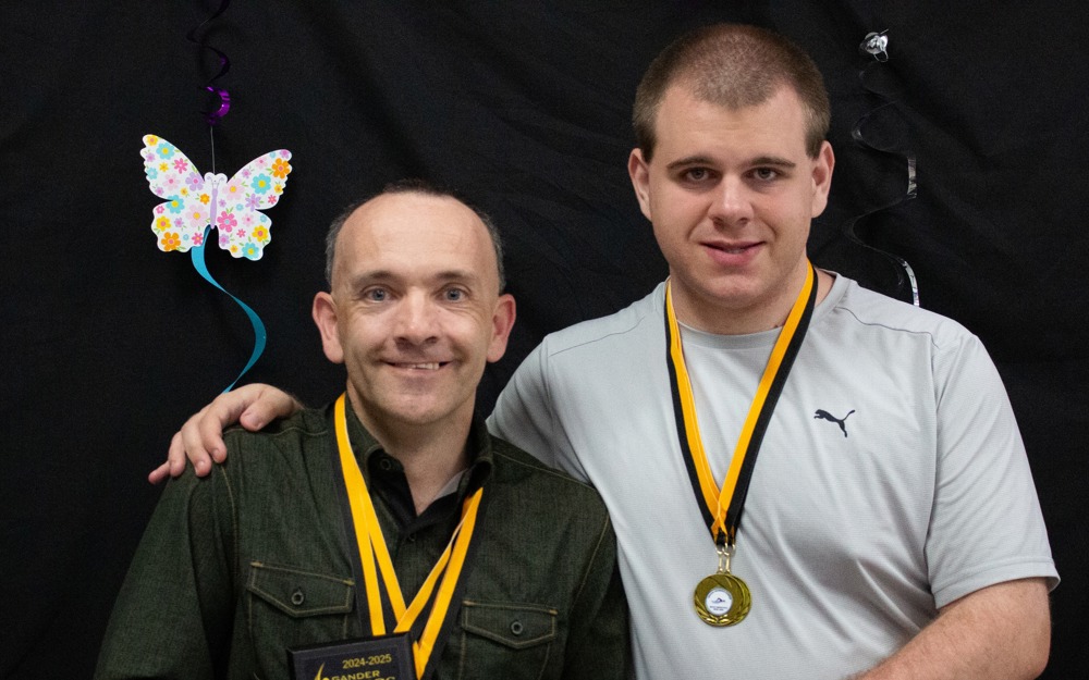 Two Special Olympics Newfoundland & Labrador athletes are smiling and posing for the camera. They both have medals hanging around their necks, and one athlete on the left is holding a plaque he won. The athlete on the right has one arm around the athlete on the left and his holding his other hand with the athlete.