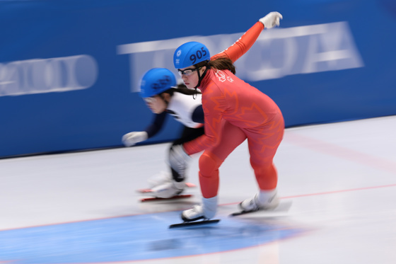 Canadian female speed skater in red skin suit. In the background is another speed skater in a black and white skin suit. 