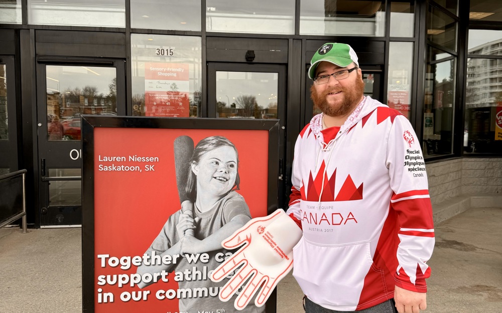 A male athlete with a red, Canada, Special Olympics Saskatchewan jersey, a green baseball cap, and a red and white foam hand, poses and smiles for the camera in front of a donation sign.