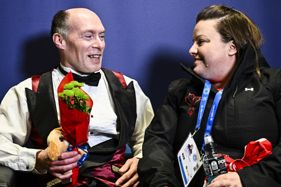 A figure skater holds a small bouquet of flowers and teddy bear while sitting and smiling with his coach.