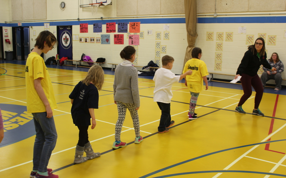 Five children in a line following their instructor. Everyone is in a gymnasium and the kids are following the lines on the floor. We can only see the children's backs as they are facing away from the camera.