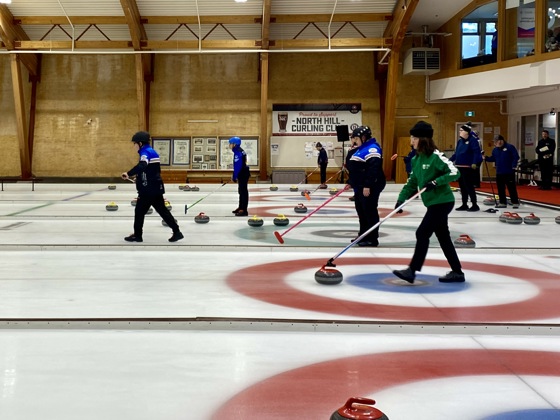A wide shot action photo of curling games in play. Multiple curling athletes can be seen with their curling sticks, getting ready to move the curling stone across the ice.