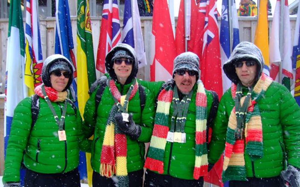 Four athletes are posing for a group photo in matching green puffer jackets and green, yellow, and red striped scarves. They are smiling for the camera as snow is falling around them. Behind them, are different flags.