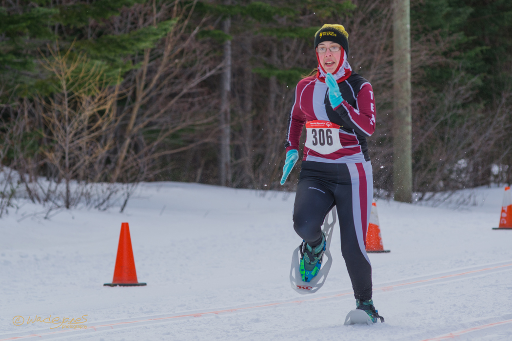 An action shot photo of a snowshoeing athlete in a competition. She is running down the track that is lined with orange pylons. She is dressed in athleisure fit for her sport and the winter weather, and has the number "306" pinned to her top.
