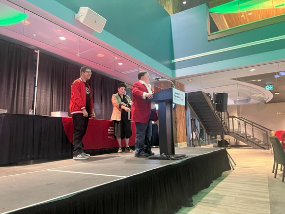 Three individuals are standing on a stage. The angle is taken from off stage looking up at them diagonally. Two individuals are standing back while one is at the podium giving a speech.