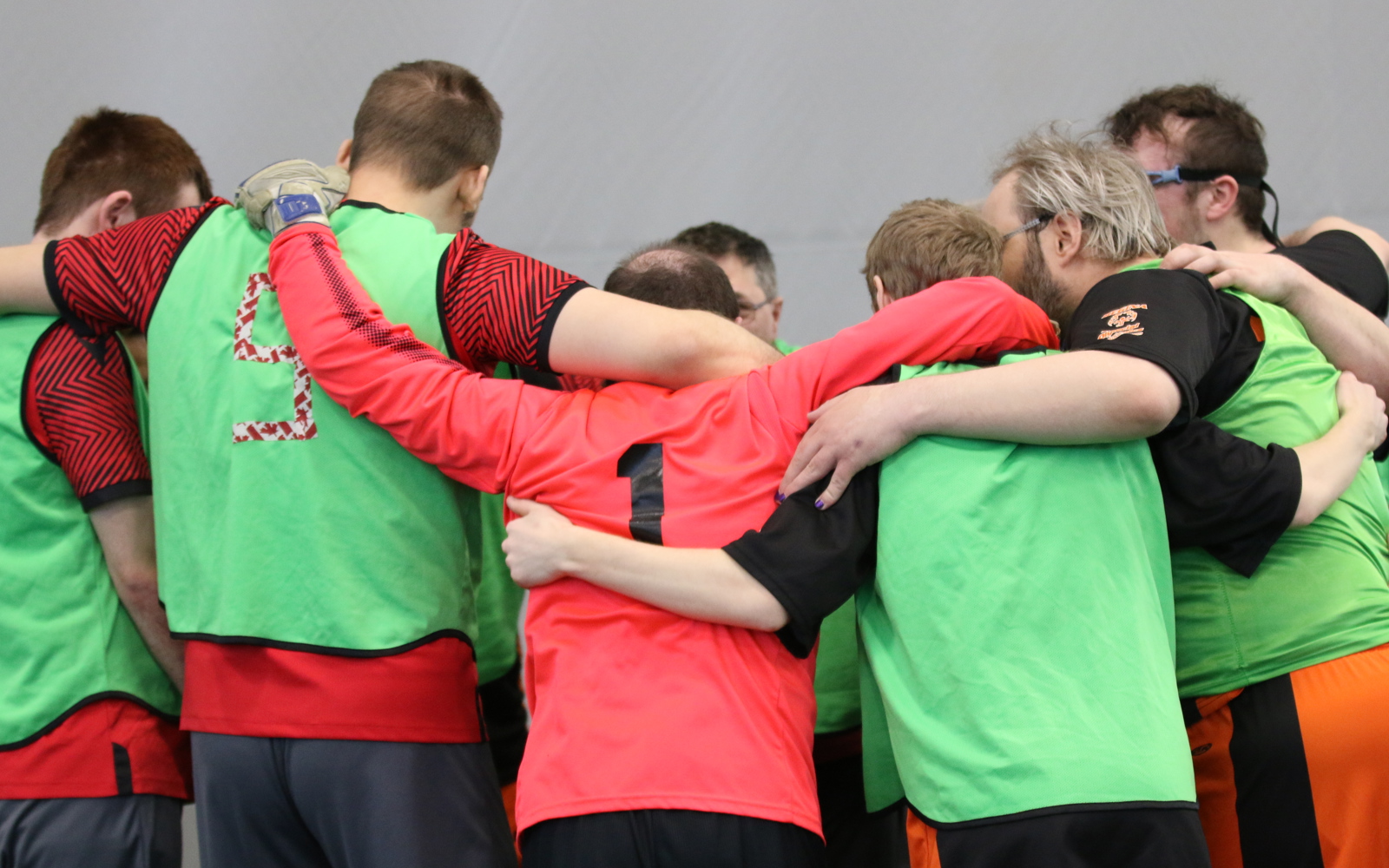Athletes on the same team are huddled together before their game. Their backs are turned to the camera. They are wearing red jerseys with green pinnies on.