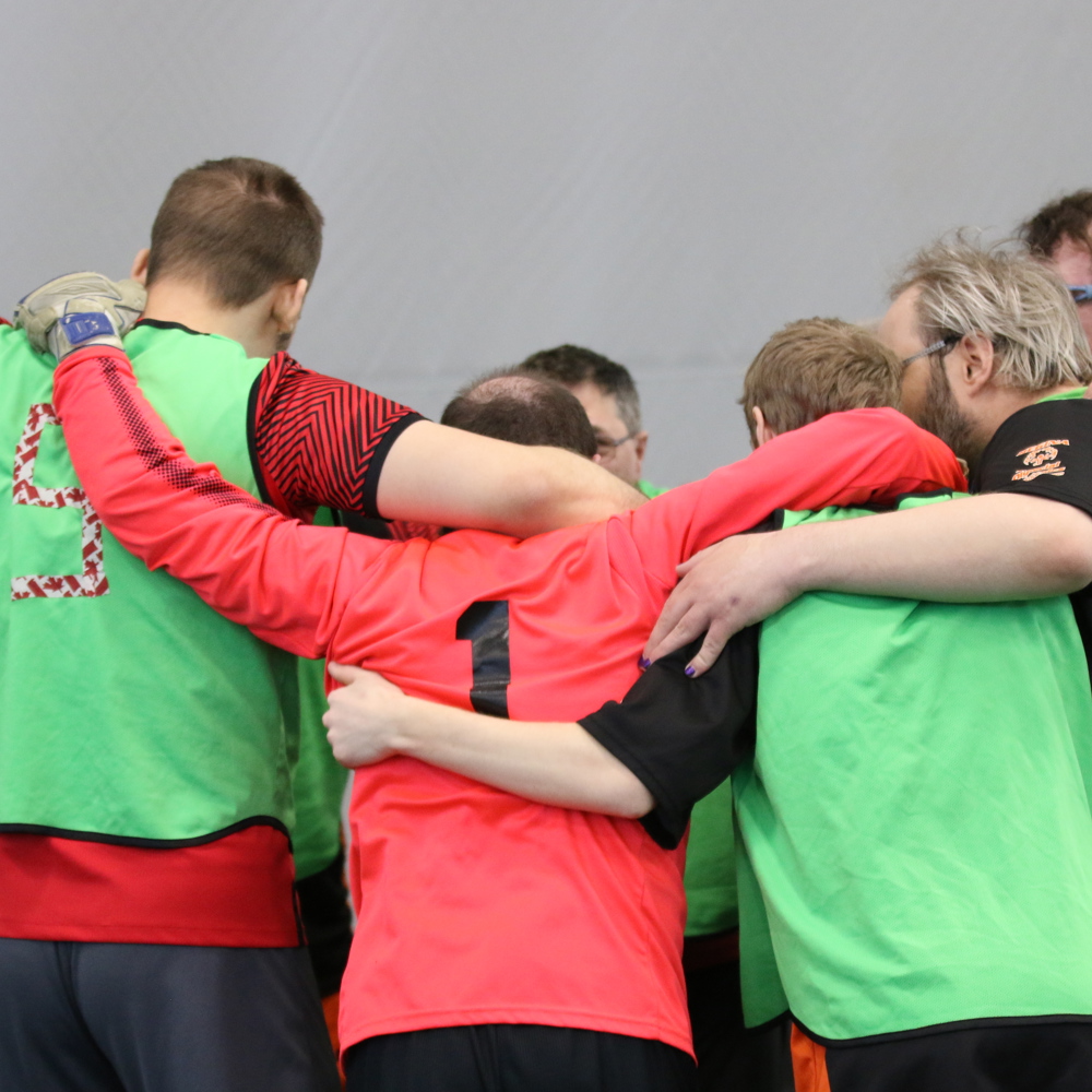 Athletes on the same team are huddled together before their game. Their backs are turned to the camera. They are wearing red jerseys with green pinnies on. 