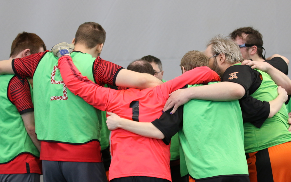 Athletes on the same team are huddled together before their game. Their backs are turned to the camera. They are wearing red jerseys with green pinnies on. 