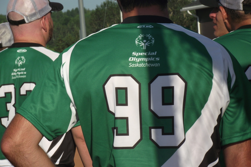 Multiple athletes are grouped together. We can only see their backs as the image is a close up of the back of an athlete's jersey. The jersey is green and has the Special Olympics Saskatchewan logo and the number 99 on it.