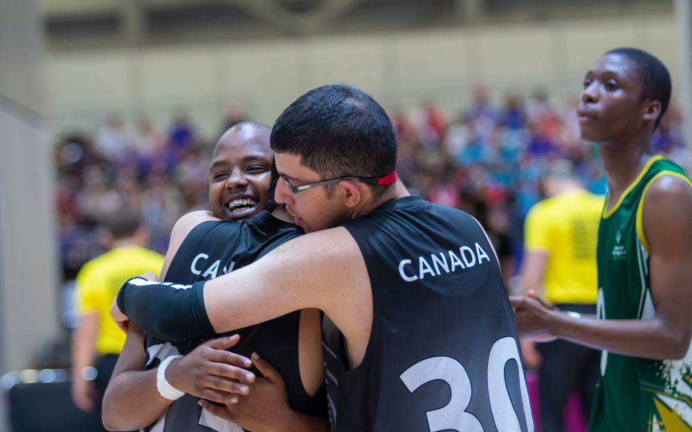 Three athletes engaging in a group hug. For two of the athletes we can only see their backs and they are wearing black "Canada" jerseys. The one athlete we can see is smiling very happily.