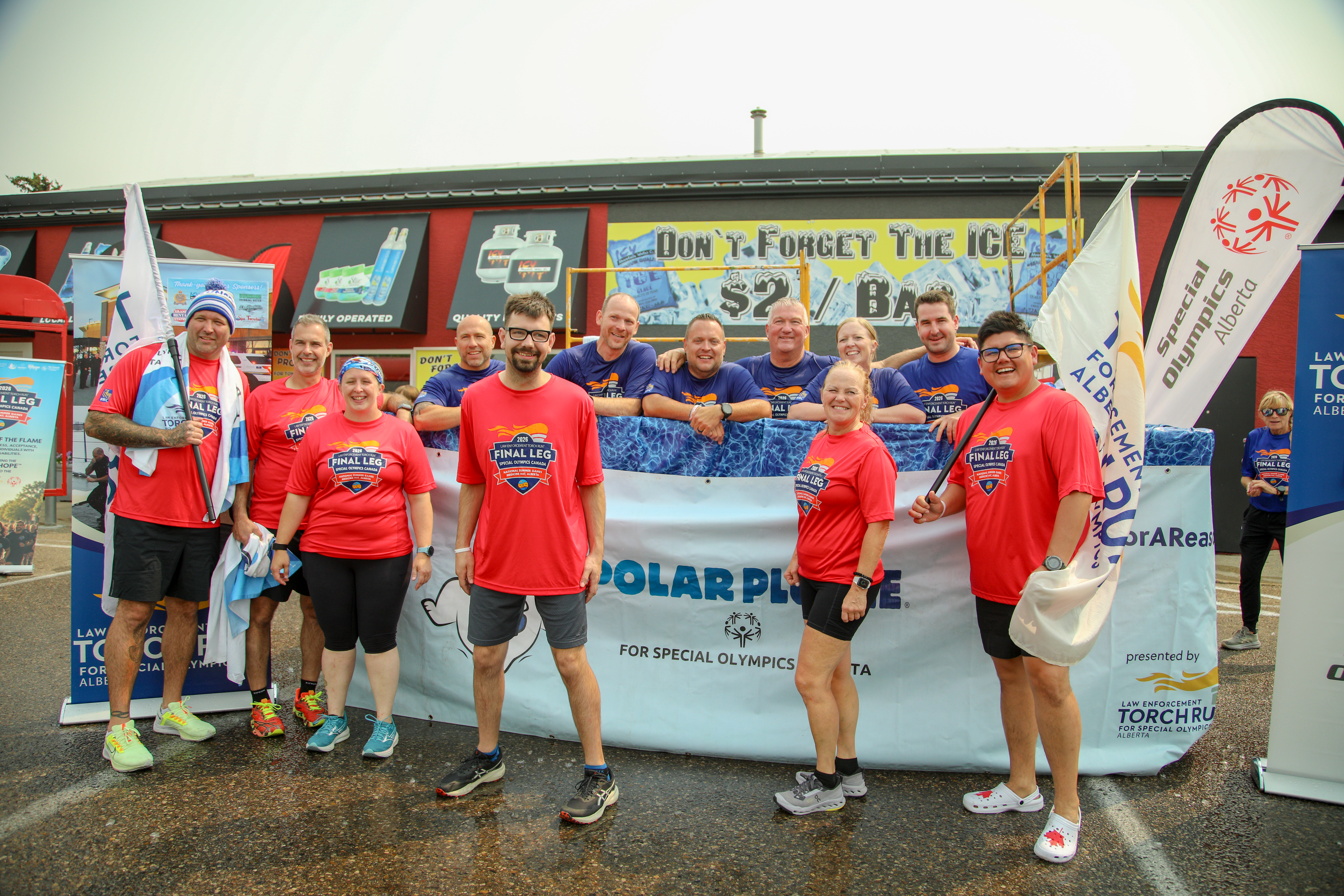 Law Enforcement Torch Run Participants standing and smiling for the camera in front of a white Polar Plunge banner. The front row of people are in matching red t-shirts while the back row of people are in matching blue t-shirts. Law Enforcement Torch Run Participants standing and smiling for the camera in front of a white Polar Plunge banner. The front row of people are in matching red t-shirts while the back row of people are in matching blue t-shirts.