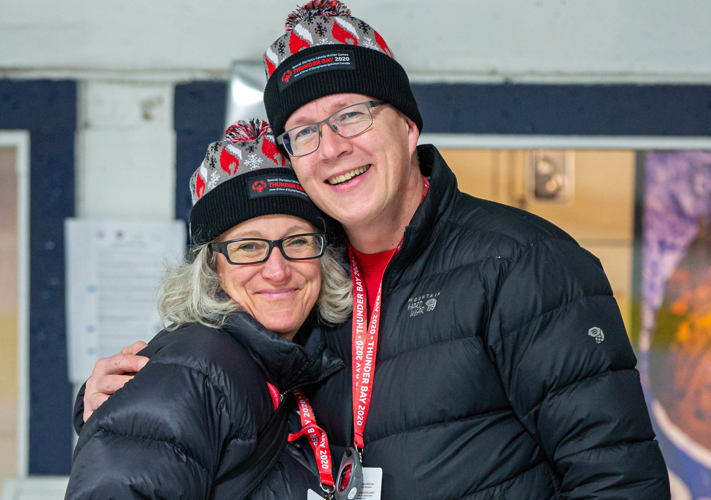 Two people in black winter jackets and Thunder Bay 2020 toques smile at the camera