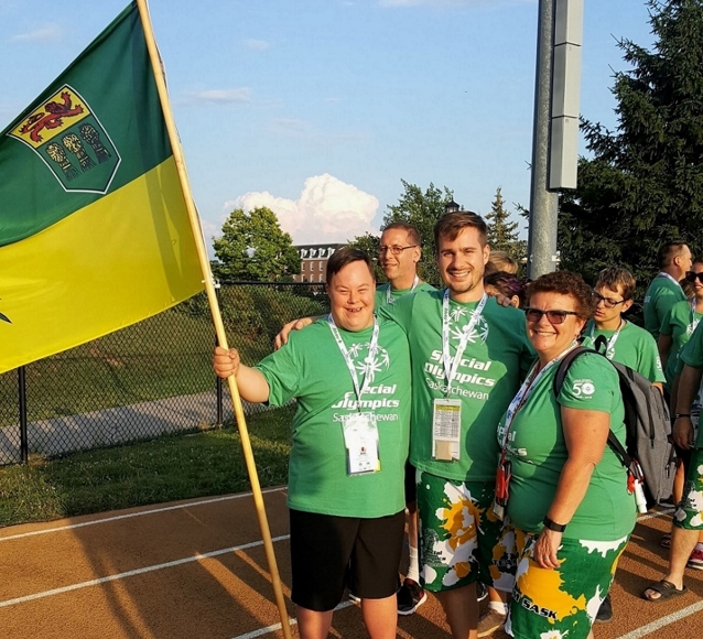 Three Special Olympics Saskatchewan athletes in green "Special Olympics Saskatchewan" t-shirts, are standing on the track. They have their arms wrapped behind each other and are smiling for the camera. One athlete, the one on left, is holding a big Saskatchewan flag.