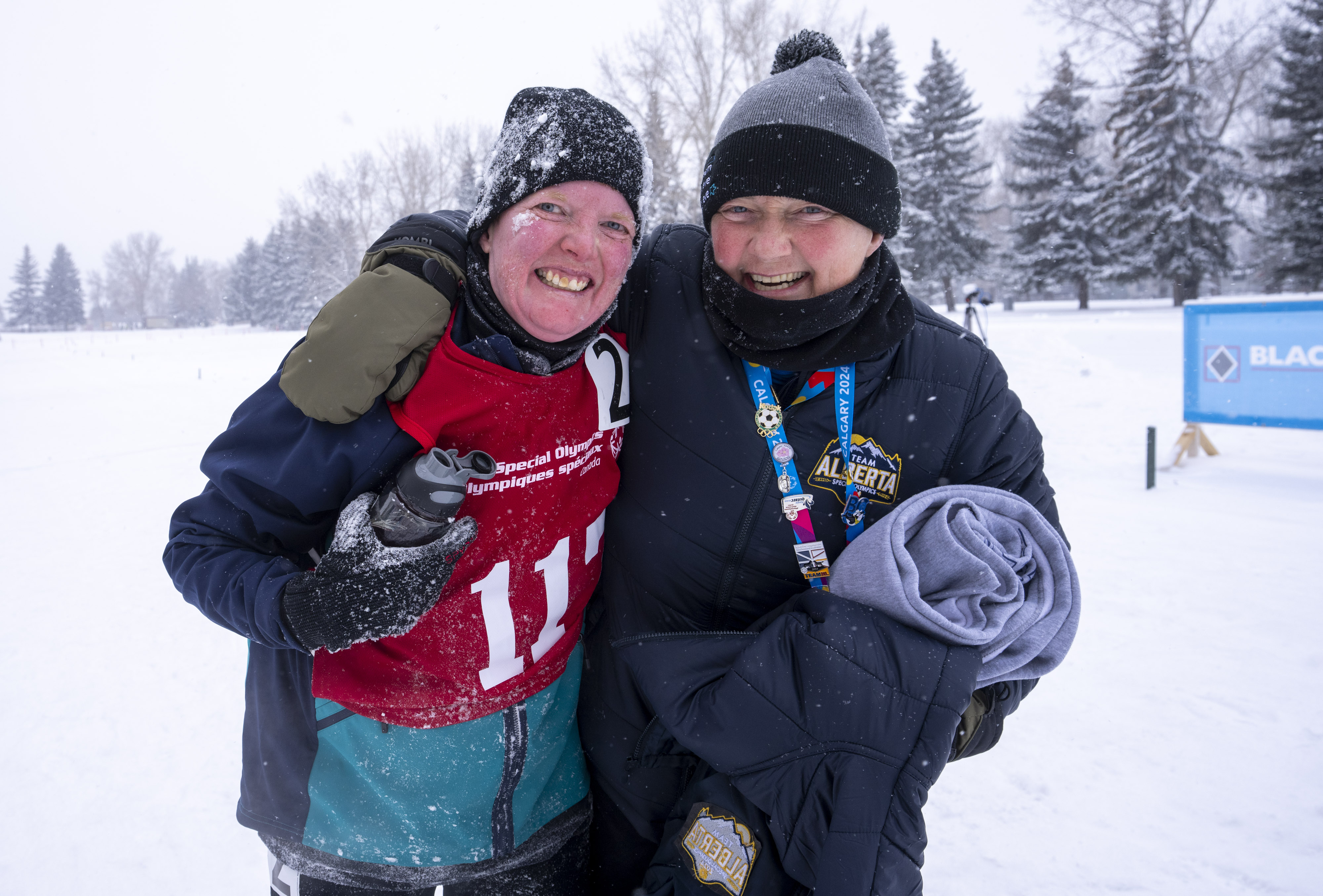 Two people with their arms around each other, smiling into the camera. One is a woman in a red pinny and one is a man with a black jacket. The background is covered in snow. Two people with their arms around each other, smiling into the camera. One is a woman in a red pinny and one is a man with a black jacket. The background is covered in snow.