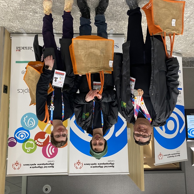 Three athletes are standing in front of Healthy Athletes banners and smiling for the camera. They are holding orange tote bags, and wearing all black with colourful lanyards around their necks.