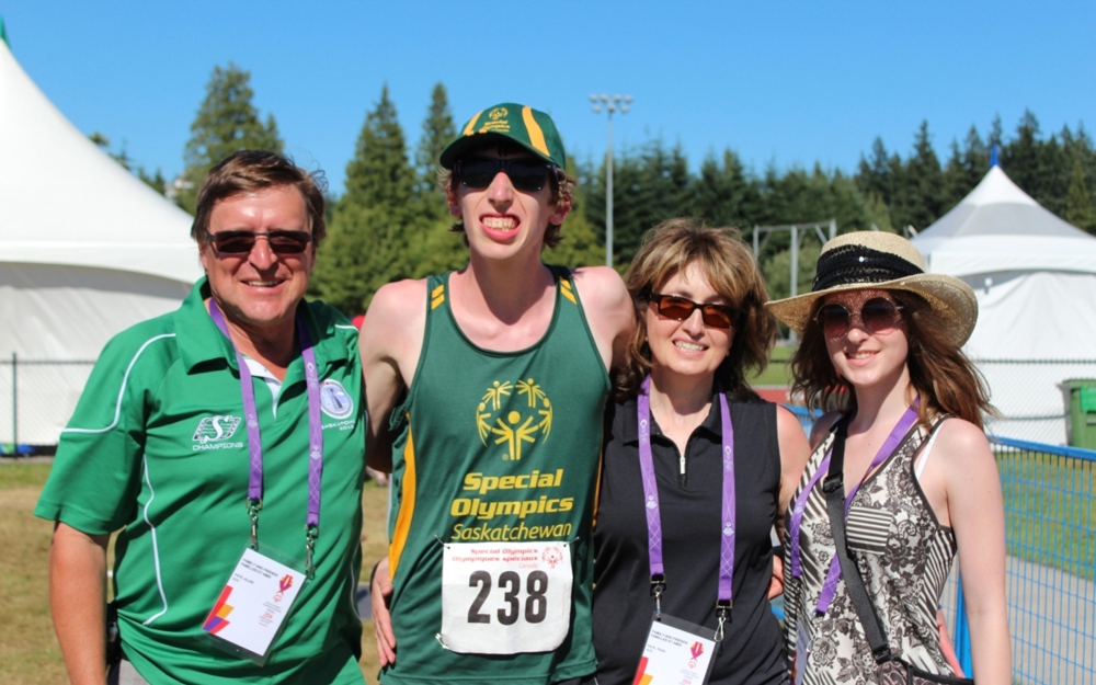 A family of four, posing for a group photo. From left to right is dad, the athlete, mom, and sister. The athlete is wearing a Special Olympics Saskatchewan tank top with the number 238 pinned to his shirt. His family members are wearing colourful lanyards to show that they are with him as supporters.