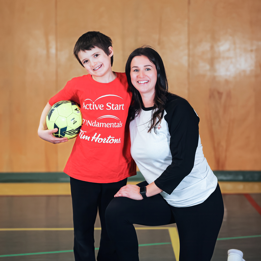 Young boy with dark hair in a red Active Start shirt and black pants holding a soccer ball. A women with long dark hair in a black and white shirt and black pants kneels beside him. Background is a gym.