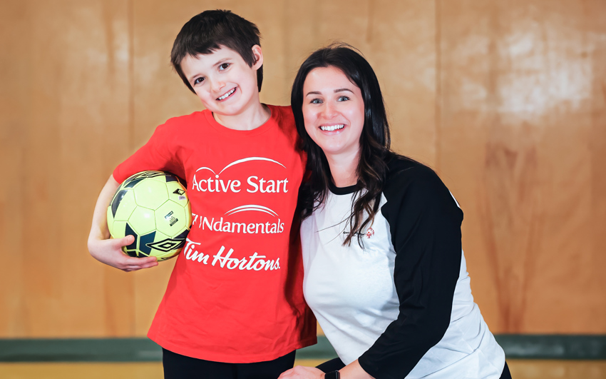 Young boy with dark hair in a red Active Start shirt and black pants holding a soccer ball. A women with long dark hair in a black and white shirt and black pants kneels beside him. Background is a gym.