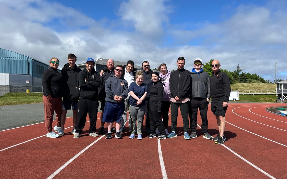 A group photo of a Special Olympics Newfoundland & Labrador team. There is 13 people in the photo and they are all standing on the track.