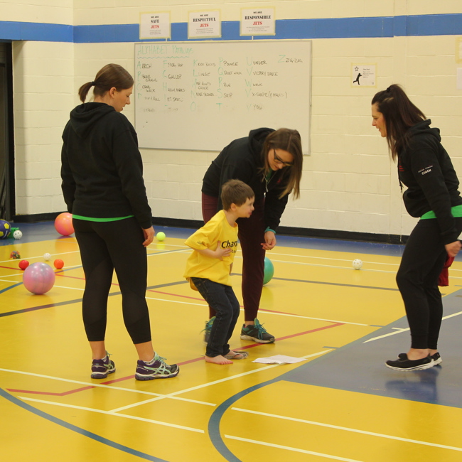 Three volunteers and coaches are standing around a young athlete, assisting him with the activity. They are all standing in a gymnasium and a variety of different sized and coloured balls can be seen in the background.