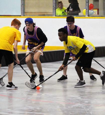 Four athletes, two in yellow shirts and two in black shirts. All have on black shorts. Each is holding a hockey stick and there is an orange ball on the floor. Four athletes, two in yellow shirts and two in black shirts. All have on black shorts. Each is holding a hockey stick and there is an orange ball on the floor.