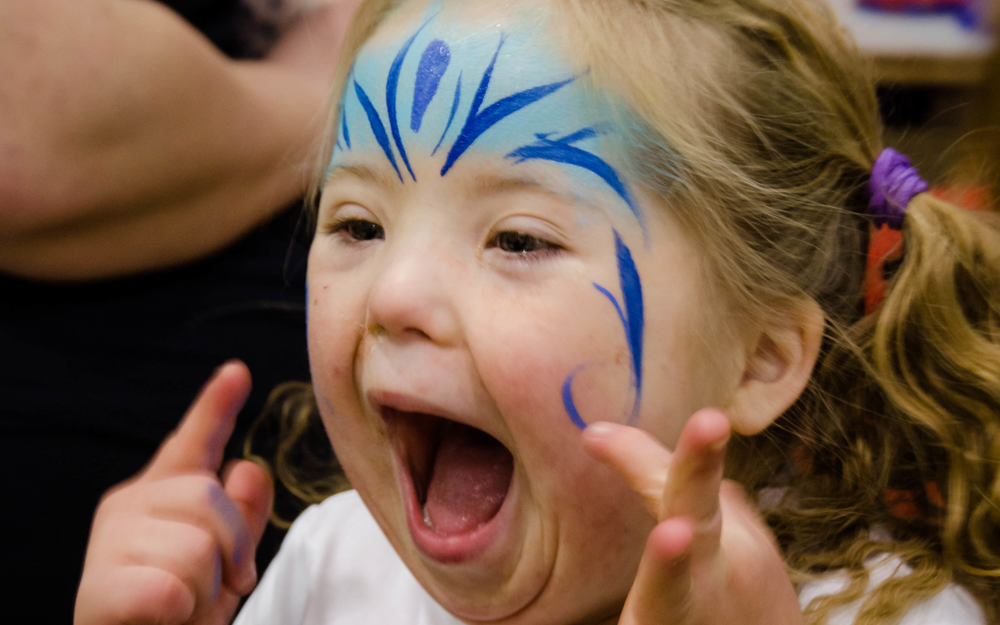 A young girl athlete has her face painted with blue paint on her forehead and cheeks, shows a very happy expression. She is turned diagonally to the left away from the camera, but you can still see her face. She is wearing a white Elsa t-shirt.
